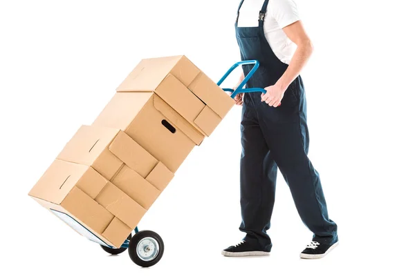 Cropped view of delivery man in overalls transporting hand truck loaded with cardboard boxes isolated on white — Stock Photo