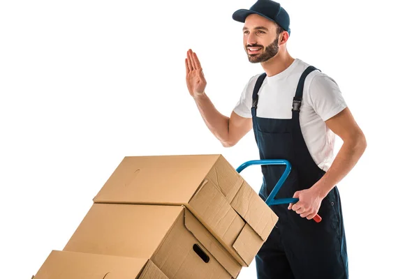 Happy delivery man transported cardboard boxes loaded on hand truck and showing hello gesture isolated on white — Stock Photo