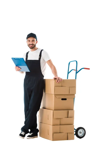 Allegro uomo di consegna in piedi vicino camion manuale caricato con scatole di cartone e tenendo appunti isolati su bianco — Foto stock