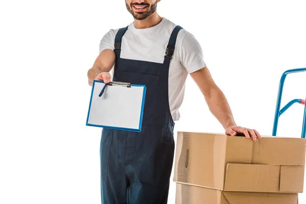 Cropped view of delivery man standing near cardboard boxes and holding clipboard with blank paper isolated on white — Stock Photo