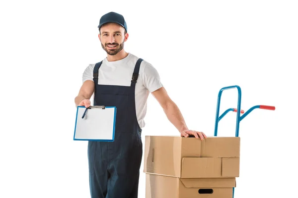 Handsome delivery man standing near cardboard boxes loaded on hand truck and holding clipboard with blank paper isolated on white — Stock Photo