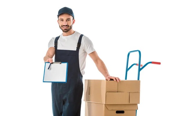 Beau livreur debout près de boîtes en carton chargé sur camion à main et tenant le presse-papiers avec du papier blanc isolé sur blanc — Photo de stock