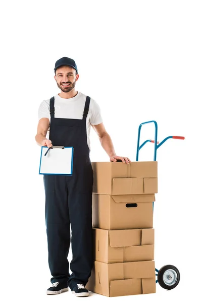 Smiling delivery man holding clipboard with blank screen while standing near hand truck loaded with cardboard boxes isolated on white — Stock Photo