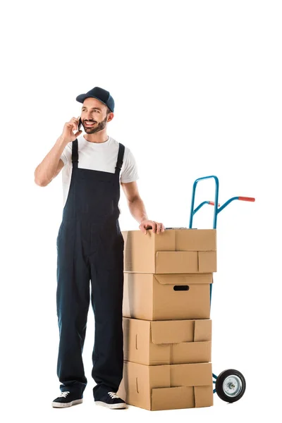 Hombre alegre de la entrega que habla en el teléfono inteligente mientras que está parado cerca del camión de mano con las cajas de cartón aisladas en blanco - foto de stock