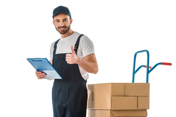 Smiling delivery man showing thumb up while standing near hand truck with carton boxes and looking at camera isolated on white — Stock Photo