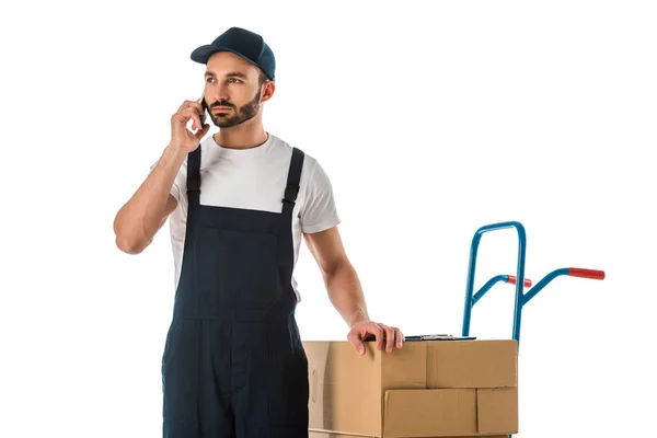 Hombre de entrega seria hablando en el teléfono inteligente mientras está de pie cerca de cajas de cartón cargadas en camión de mano aislado en blanco - foto de stock