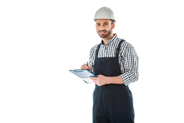 Sonriente trabajador de la construcción escribiendo en el portapapeles y mirando a la cámara aislada en blanco - foto de stock