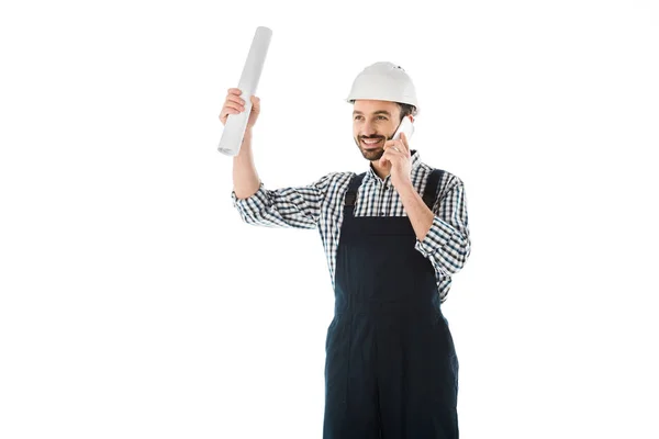 Smiling construction worker talking on smartphone while holding rolled paper in raised hand isolated on white — Stock Photo