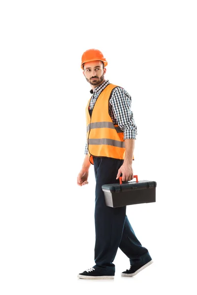 Серьезный строительный работник в бронежилете и шлеме, держащий ящик с инструментами и оглядывающийся назад изолированный на белый — стоковое фото