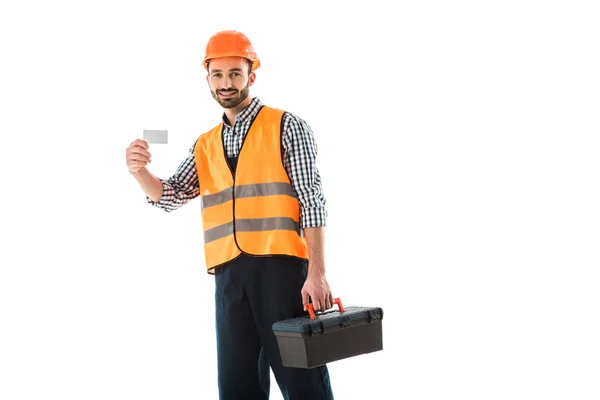 Alegre trabajador de la construcción con caja de herramientas con tarjeta de visita en blanco y mirando a la cámara aislada en blanco - foto de stock