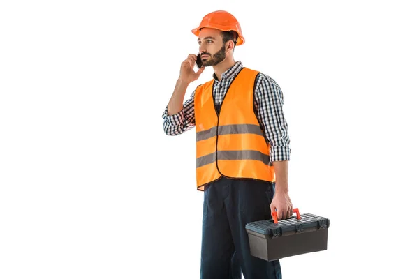 Trabajador de la construcción atento hablando en el teléfono inteligente mientras sostiene la caja de herramientas y mirando hacia otro lado aislado en blanco - foto de stock