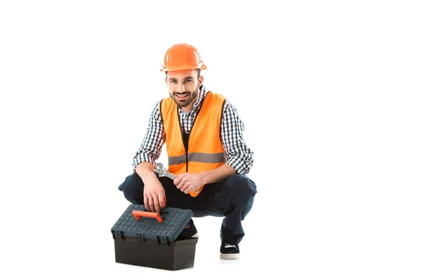 Alegre trabajador de la construcción sentado cerca de la caja de herramientas y la celebración de llave ajustable aislado en blanco — Stock Photo