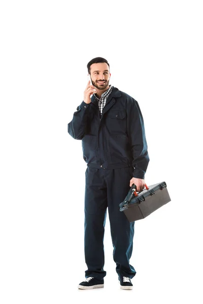 Smiling workman in overalls talking on smartphone and holding toolbox isolated on white — Stock Photo