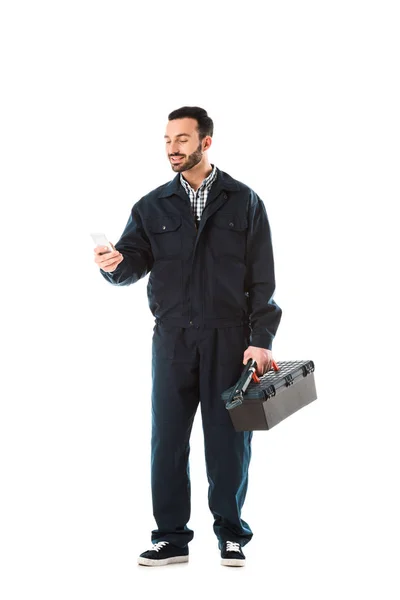 Obrero alegre en overoles usando teléfono inteligente y sosteniendo la caja de herramientas aislada en blanco - foto de stock