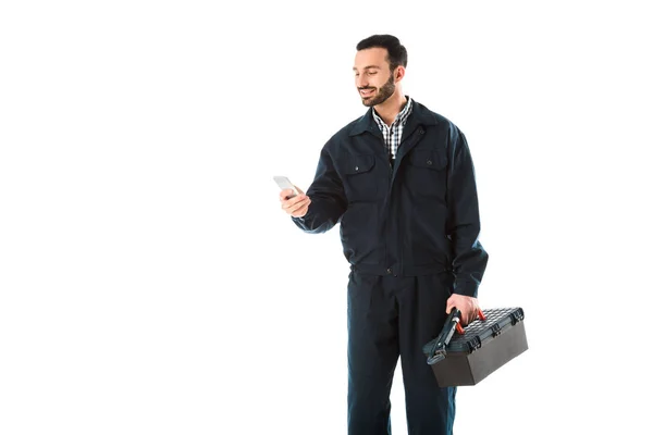 Smiling workman in overalls using smartphone while holding toolbox isolated on white — Stock Photo