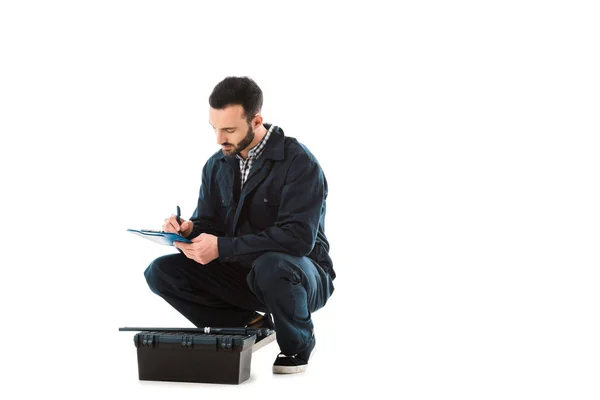 Attentive workman writing on clipboard while sitting near toolbox isolated on white — Stock Photo