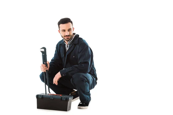 Obrero sonriente sosteniendo llave ajustable mientras está sentado cerca de la caja de herramientas aislado en blanco - foto de stock