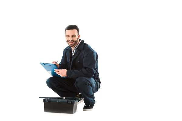 Cheerful workman writing on clipboard while sitting near toolbox and looking at camera isolated on white — Stock Photo