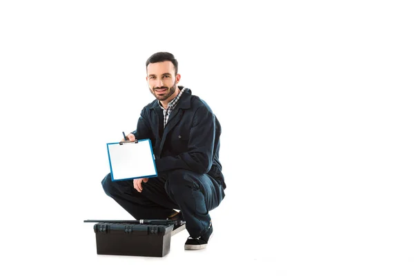 Fontanero sonriente guapo sosteniendo portapapeles con papel en blanco mientras está sentado cerca de la caja de herramientas aislado en blanco - foto de stock