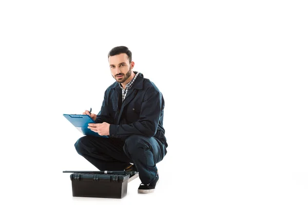 Reparador serio escribiendo en el portapapeles mientras está sentado cerca de la caja de herramientas y mirando la cámara aislada en blanco - foto de stock