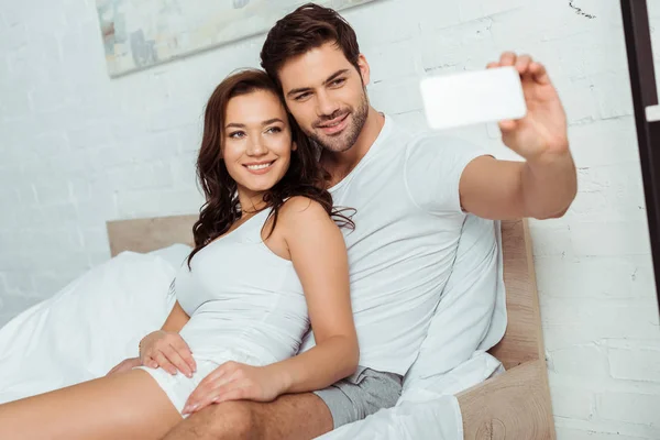 Foco seletivo de homem feliz tomando selfie com namorada atraente no quarto — Fotografia de Stock