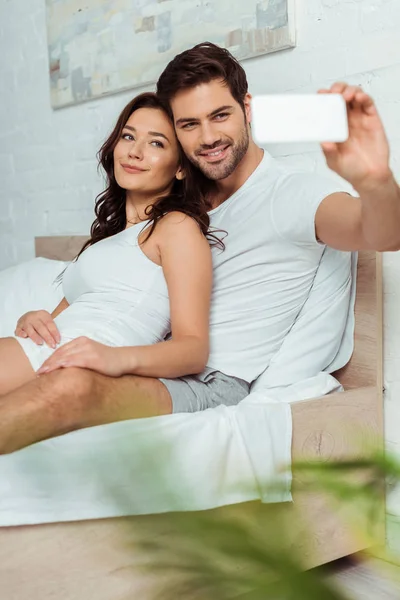 Enfoque selectivo de hombre feliz tomando selfie con novia alegre en el dormitorio - foto de stock