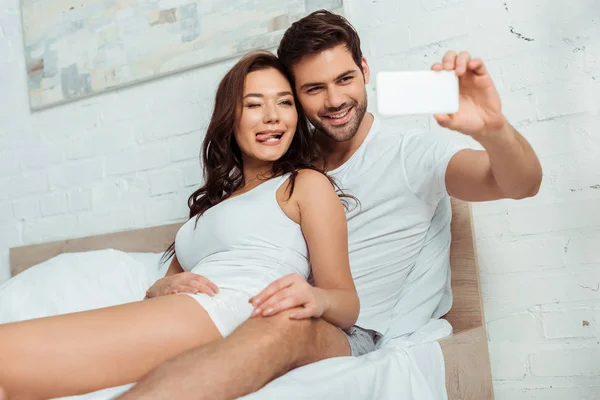 Selective focus of happy man taking selfie with girlfriend showing tongue in bedroom — Stock Photo
