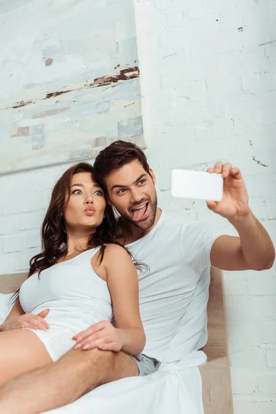 Happy man showing tongue near attractive girl with duck face while taking selfie — Stock Photo