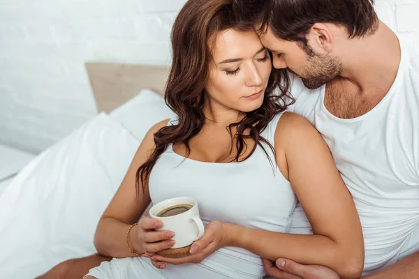 Bell'uomo vicino alla donna attraente che tiene una tazza di caffè a letto — Foto stock