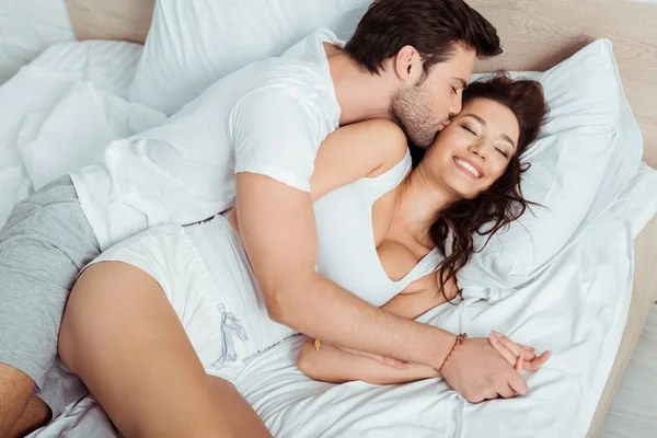 Вид зверху щасливого чоловіка цілує щоку веселої молодої жінки, лежачи на ліжку з закритими очима — стокове фото