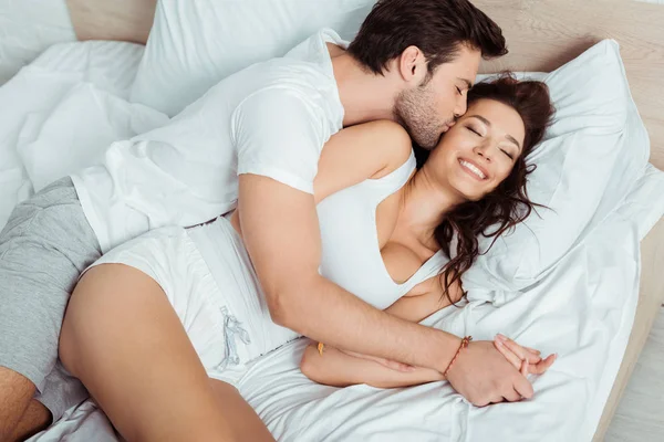 Vista aérea del hombre feliz besando mejilla de la joven alegre acostada en la cama con los ojos cerrados — Stock Photo