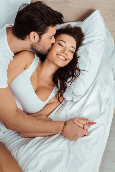 Вид зверху чоловіка цілує щоку веселої молодої жінки, лежачи на ліжку з закритими очима — стокове фото