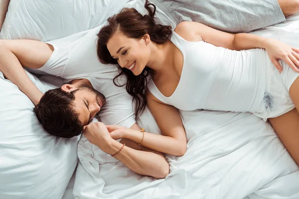 Vista superior da mulher alegre olhando para o homem, mantendo as mãos e deitado na cama — Fotografia de Stock