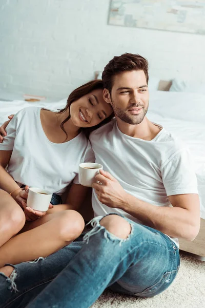 Hombre guapo y mujer alegre sosteniendo tazas en el dormitorio - foto de stock
