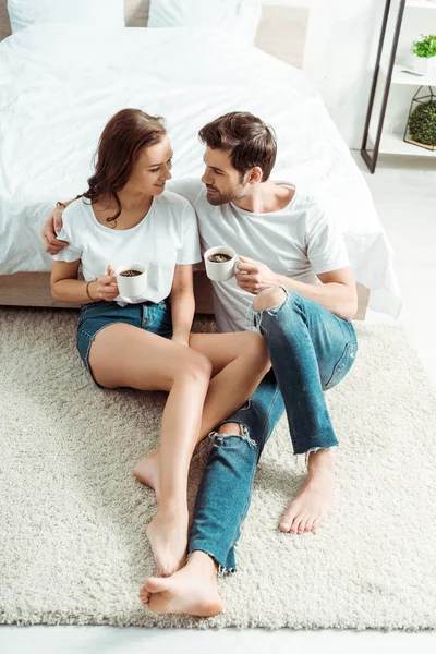 Vista aérea del hombre guapo sentado con chica alegre en la alfombra y la celebración de la taza en el dormitorio - foto de stock