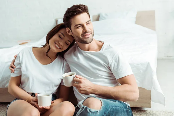 Hombre feliz sentado con chica alegre y sosteniendo la taza en el dormitorio - foto de stock