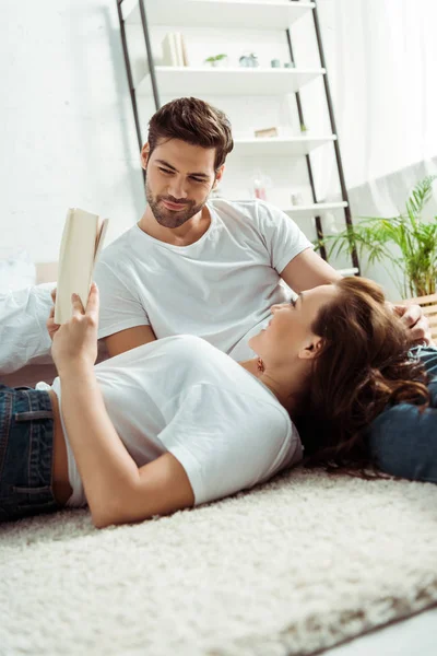 Low angle view of handsome man looking at book in hands of girlfriend lying on carpet in bedroom — Stock Photo