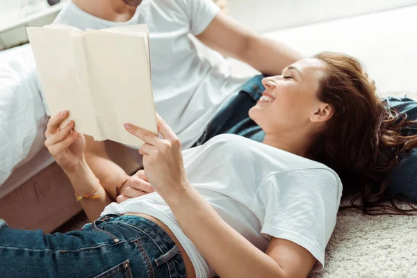 Обрізаний вид чоловіка біля веселої дівчини тримає книгу, лежачи на килимі в спальні — стокове фото