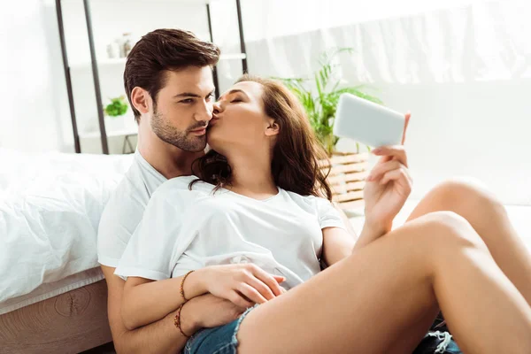 Привлекательная девушка целует щеку мужчины, делая селфи на смартфоне — стоковое фото
