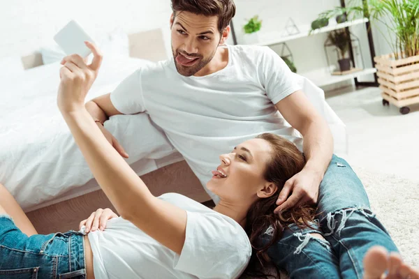 Enfoque selectivo de la niña y el hombre mostrando lenguas mientras toma selfie en el dormitorio - foto de stock