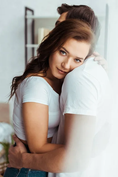 Novio abrazando atractiva novia mirando a la cámara en casa - foto de stock