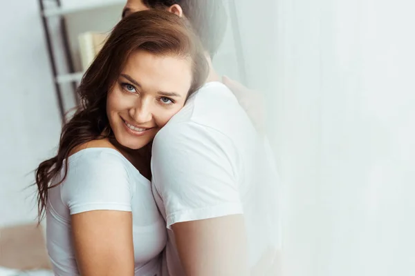 Hombre abrazando atractiva y alegre novia mirando a la cámara en casa - foto de stock