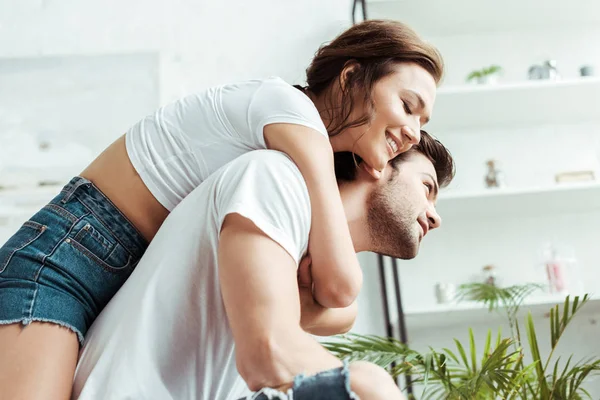 Feliz chica abrazando guapo novio en blanco camiseta y sonriendo en casa - foto de stock