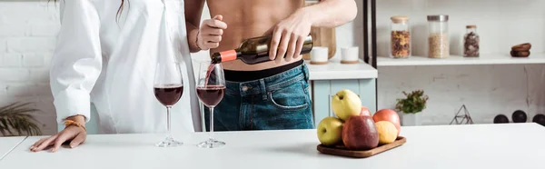 Панорамний знімок без сорочки, що заливає червоне вино в келихах для вина біля дівчини на кухні — стокове фото