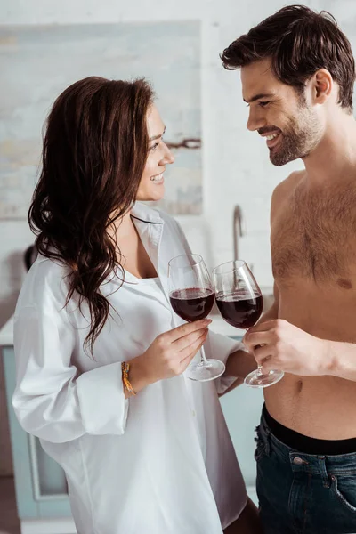 Hombre musculoso alegre sosteniendo copa de vino cerca de chica atractiva y feliz en casa - foto de stock