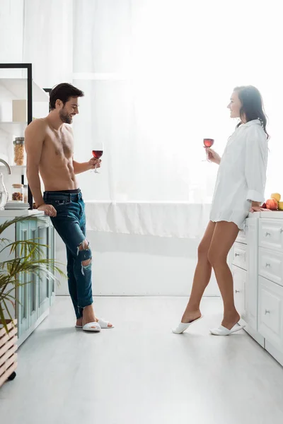 Hombre sin camisa guapo y atractiva chica morena sosteniendo copas de vino en la cocina moderna - foto de stock