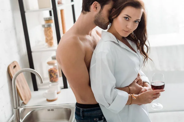 Homem muscular abraçando atraente morena menina segurando copo de vinho e olhando para a câmera — Fotografia de Stock