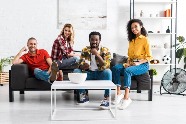 Happy smiling multiethnic friends sitting on couch together near table with popcorn in bowl — Stock Photo