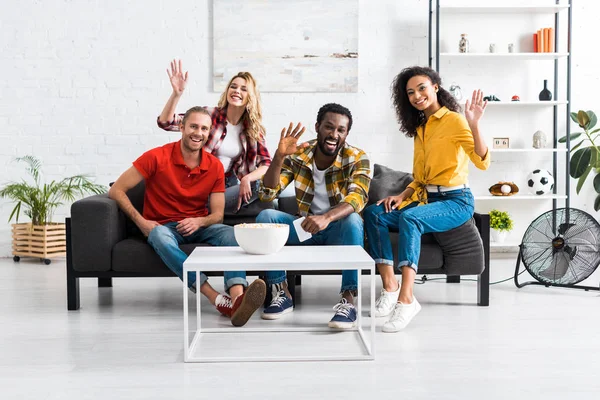 Amigos multiculturales sonrientes están sentados en el sofá, saludando con las manos y divirtiéndose juntos - foto de stock
