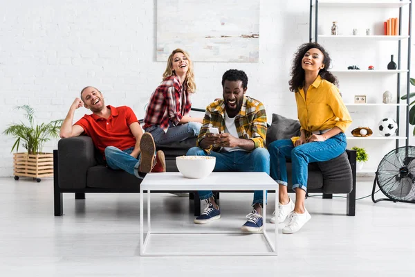 Jeunes hommes et femmes multiculturels heureux et excités assis sur le canapé et s'amusant ensemble — Photo de stock